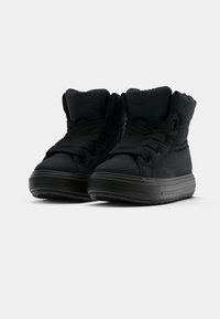 Et par svarte polstrede høye sneakers med tjukke gummisåler og brede svarte lisser på en hvit bakgrunn.