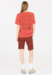 Punainen puuvillainen T-paita, jossa on graafinen printti selässä, yhdistettynä ruskeisiin shortseihin. Ominaisuuksiin kuuluu rento mitoitus ja pyöreä kaula-aukko.