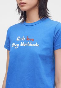 Γυναίκα με μπλε μπλουζάκι με το κείμενο "Girls love Obey Worldwide" σε λευκά και κόκκινα γράμματα μπροστά.
