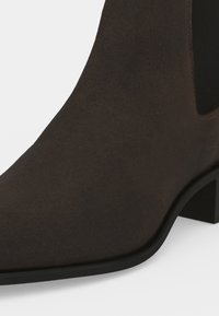 Gros plan d’une botte cheville en daim marron avec un panneau latéral élastique noir et un petit talon bloc.