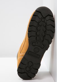 Buty Timberland z brązową zamszową cholewą, czarną gumową podeszwą z bieżnikiem i logo oraz białymi szwami wzdłuż szwów.