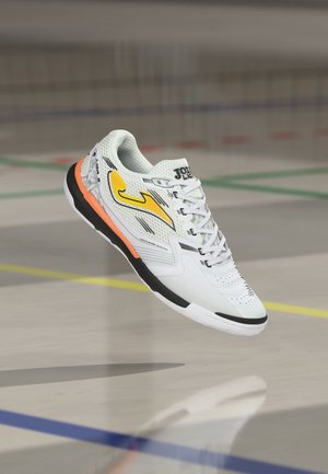 Weißer Indoorsportschuh mit gelbem Logo, schwarzen und orangen Sohlenakzenten, geschnürten Schnürsenkeln, schwebend über dem Hallenboden mit farbigen Spielfeldlinien.