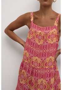 Pink sleeveless dress featuring an intricate yellow and pink floral pattern, with a square neckline and a tiered hem. Smooth fabric texture.