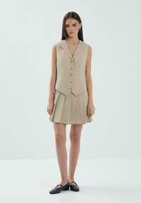 Beżowa bezrękawnik z wyciętym kołnierzem, z rzędem guzików i dopasowaną talią, w połączeniu z pasującą plisowaną spódnicą.