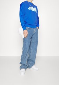 Blå Jordan-tröja med vitt logo, löst sittande denimjeans och vita sneakers. Tröjan har långa, kontrasterande ärmar.