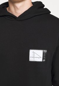Homme portant un sweat à capuche noir avec un patch blanc affichant un diagramme de triangle rectangle et le texte "URBAN" et "GRAVITY DEFYING" sur la poitrine.