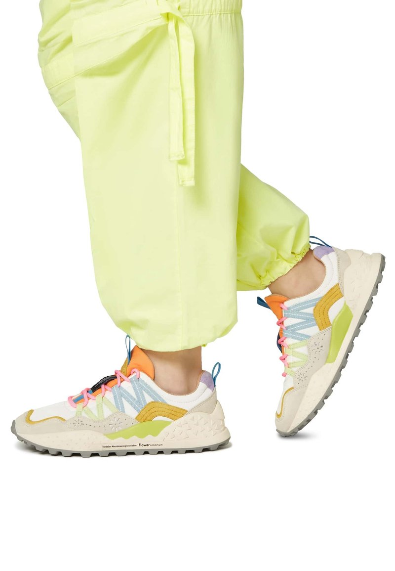 Multicolored sneakers with a mix of cream, yellow, pastel blue, and orange accents. Textured sole and laces in pink. Worn with bright yellow pants.