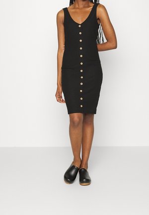 Osoba ubrana w czarną, bezrękawnikową sukienkę do kolan z guzikami z przodu oraz czarne klapki wsuwane, stojąca na tle jednolitej białej ściany.