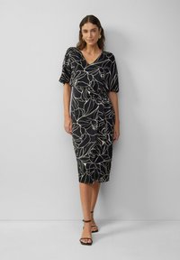 Robe noire avec un motif floral beige, décolleté en V, manches courtes et un détail de taille nouée. Tissu lisse avec une longueur au genou. Sandales simples à lanières.