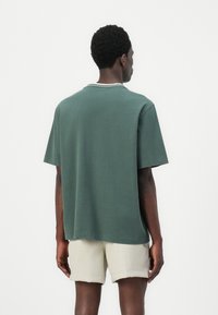 Dark green cotton T-shirt with a relaxed fit, short sleeves, and a crew neckline featuring a subtle white trim. Worn with light beige shorts.