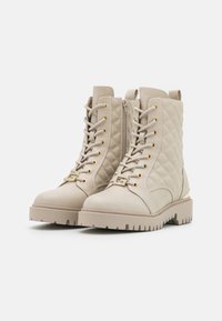 Bottes en beige à lacets avec des accents matelassés, des garnitures dorées, un col rembourré et des semelles en caoutchouc épaises. Fermeture éclair sur le côté pour un accès facile.