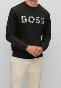 Svart sweatshirt med "BOSS" i texturerade, mönstrade bokstäver. Ribbad mudd i ärmar och nederkant. Avslappnad passform. Ljust färgade byxor.