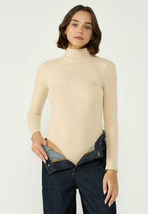 Nude long-sleeve bodysuit with high neckline and fitted design, paired with unbuttoned dark denim jeans revealing a light lining.