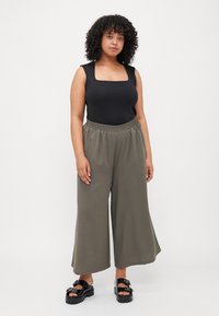 Μαύρο κορμάκι με τετράγωνη λαιμόκοψη, χαλαρές ελαιόχρωμες παντελόνες καμπάνα και μαύρες πλατφόρμες με διακόσμηση. Φυσικά σγουρά μαλλιά.