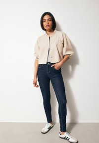 Chaqueta corta beige con un bolsillo con cremallera, camiseta gris, pantalones pitillo azul oscuro y zapatillas blancas con acentos en negro y azul.