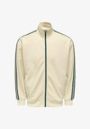 Kermanvärinen vetoketjullinen juoksutakki, jossa on tummanvihreä vetoketju ja kolme tummanvihreää raitaa, jotka kulkevat hihojen päästä olkapäähän.