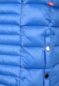 Blå puffjacka med quiltat mönster, glänsande nylontyg och silverfärgade knapppdetaljer. Har en röd varumärkesetikett på sömmen.