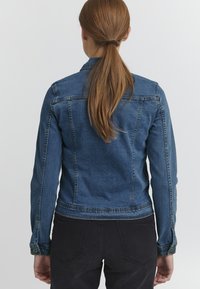 Denim jakna v temno modri barvi, z opremljenim dizajnom, krojenimi šivi in gumbastimi manšetami. Teksturirano blago in strukturirana silhueta.