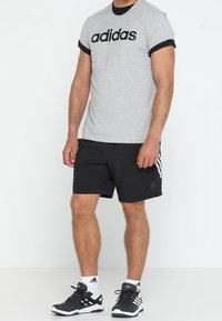 Šedé bavlněné tričko s černým lemem a velkým logem "adidas", spárované s černými kraťasy se třemi bílými pruhy a černými sportovními botami.