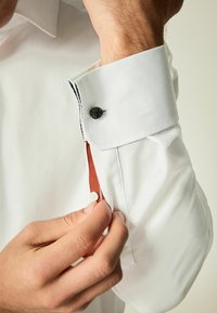 Vit skjort med texturerat tyg, med en svart knappdetalj, kontrasterande röd tyg inuti manschetten och en plastknapp.