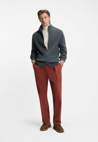 Grå stickad zip-up tröja med hög krage, parat med rostfärgade byxor och bruna snörskor. Enfärgad bakgrund.