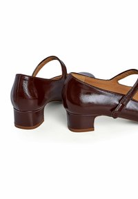 Scarpe in pelle verniciata bordeaux con tacco basso a blocco, caratterizzate da un'apertura sul retro e un delicato design a strisce incrociate. Texture liscia, forma arrotondata.