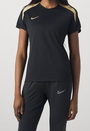 Personne portant une tenue de sport Nike noire avec des accents dorés sur les épaules et un logo Nike blanc sur le pantalon, debout devant un fond uni.
