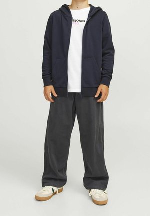 Kind draagt marineblauwe hoodie met rits, witte bedrukte T-shirt, losse zwarte broek en witte sneakers met gumzolen, staand tegen een effen achtergrond.