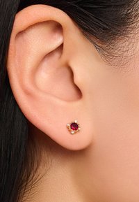 Goldene Ohrstecker mit einem zentralen roten Edelstein, der von vier kleinen weißen Akzenten umgeben ist. Nahaufnahme, die das Design und die Details hervorhebt.