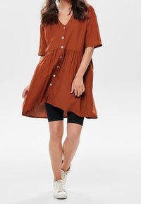 Rustfarbenes Hemdkleid mit V-Ausschnitt, kurzen Ärmeln, Knopfverschluss und gerafftem Taillengürtel; kombiniert mit schwarzen Fahrradhosen und weißen Turnschuhen.