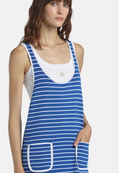 Femme portant une robe sans manches à rayures bleues et blanches par-dessus un débardeur blanc, main dans la poche droite, regardant légèrement à gauche.