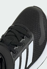 Scarpa sportiva nera con parte superiore in rete, chiusura con lacci e tre strisce bianche. La suola è testurizzata e presenta una forma arrotondata per il comfort.