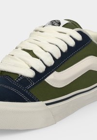 Baskets basses avec lacets blancs, panneaux en tissu bleu marine et vert olive, semelle en caoutchouc blanche et logo Vans sur la languette.