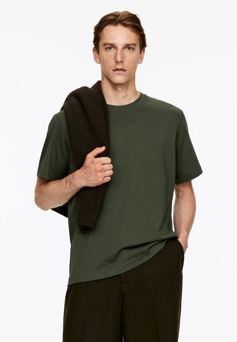 T-shirt en coton vert foncé avec un col rond, porté avec un pull texturé sombre drapé sur l'épaule, associé à un pantalon noir.