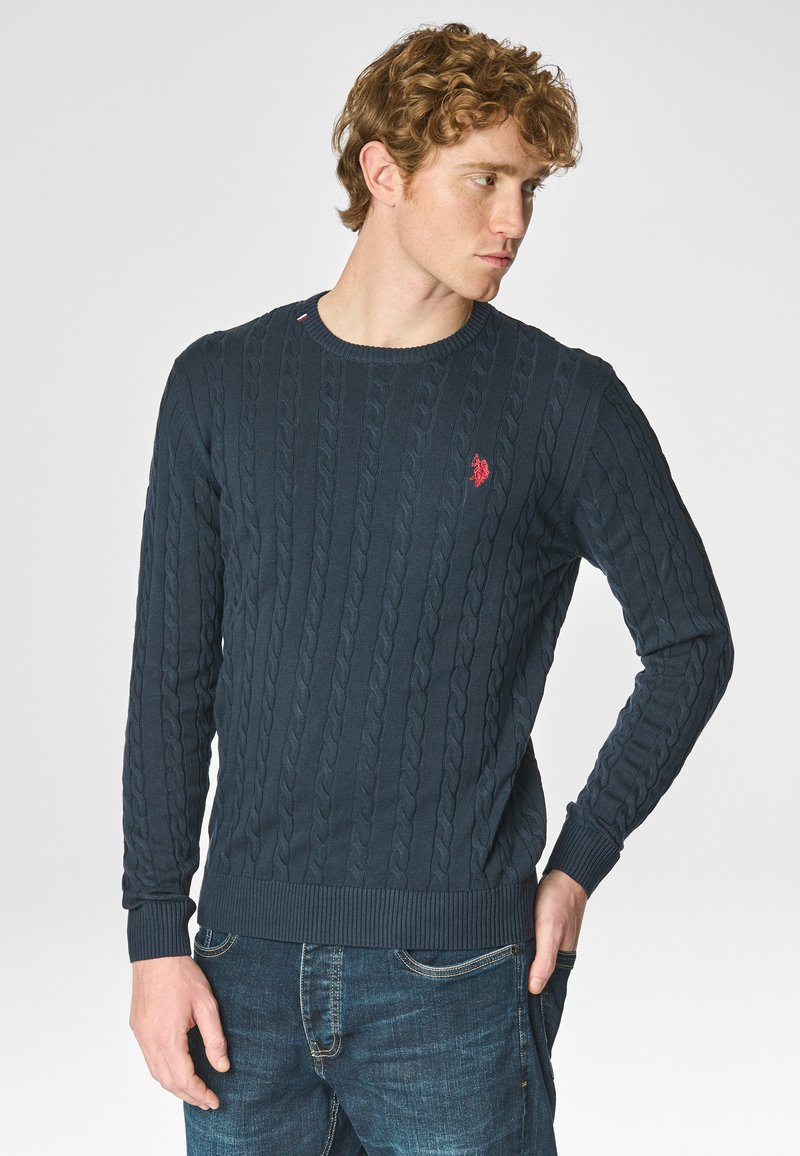 Maglione lavorato a maglia blu navy con un motivo a trecce, polsini e orlo a coste, con un piccolo logo rosso sul petto. Indossato sopra jeans in denim.