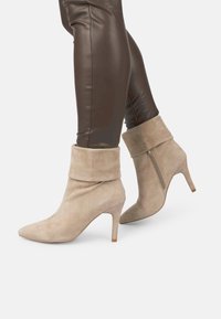 Botas de tobillo de ante color beige con puño doblado, punta afilada y tacón delgado. Cuenta con una cremallera lateral para facilitar su uso.
