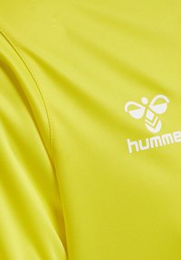 Tissu de sport jaune avec coutures visibles, un logo d'abeille stylisé blanc et le nom de la marque « hummel » imprimés sur la surface.