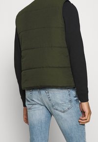 Dunkelgrüne Steppweste mit horizontalen Nähten, getragen über einem schwarzen langärmeligen Shirt, kombiniert mit hellblauen Jeans.