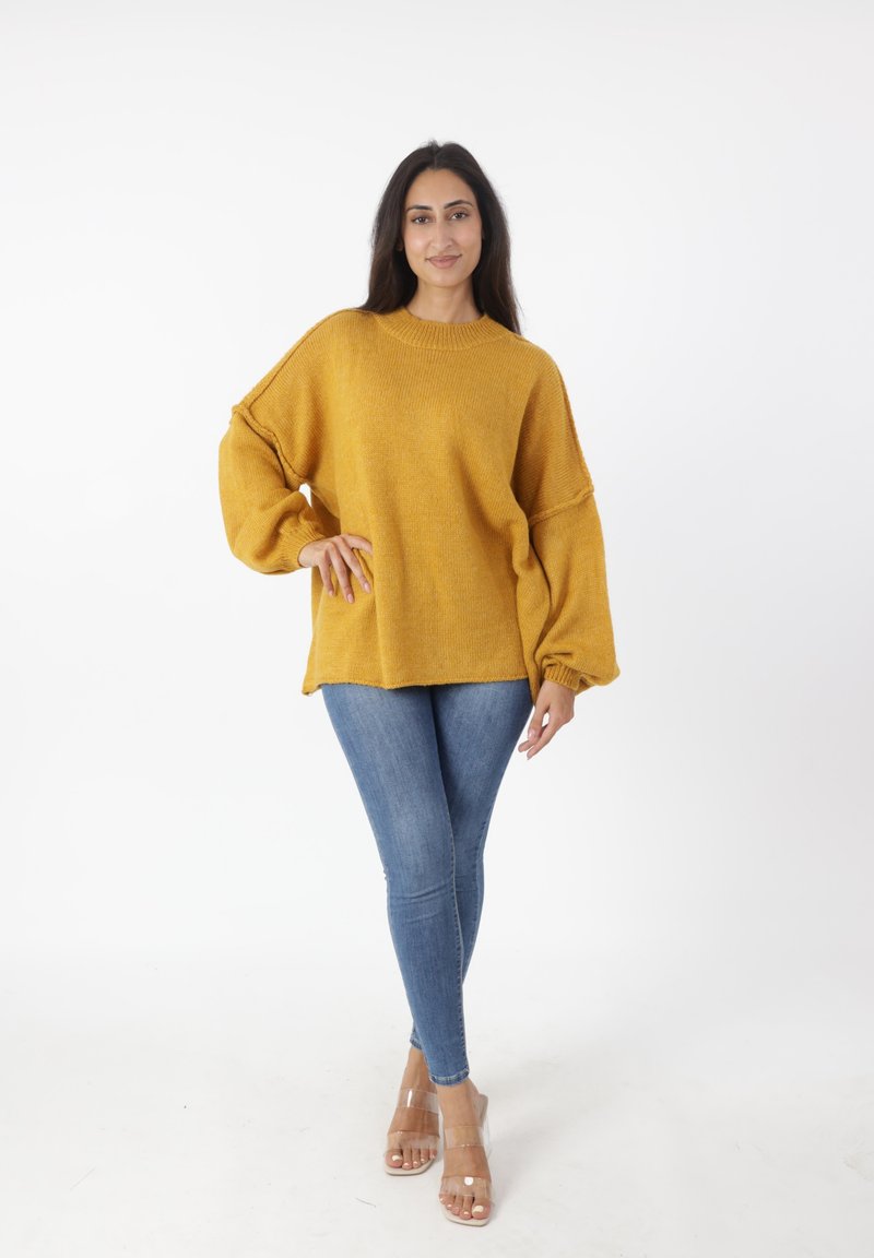 Musztardowy, oversize'owy sweter z dzianiny o balonowych rękawach, w zestawieniu z przylegającymi niebieskimi dżinsami i przezroczystymi sandałami na słupku.