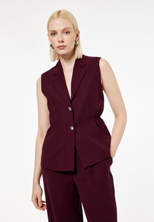 Blonde vrouw die een mouwloos donkerbordeaux getailleerd vest draagt met zilveren knopen en bijpassende wijde broek tegen een effen achtergrond.