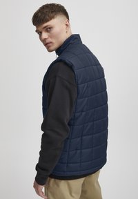 Marinblå vadderad väst med fyrkantig quiltning, hög krage och ärmlös design, lager över en svart långärmad tröja.