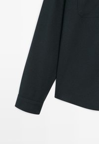 Dettaglio di una camicia nera a maniche lunghe che mostra un tessuto testurizzato e un polsino, con una leggera curvatura dell'orlo e una tasca posteriore delineata.