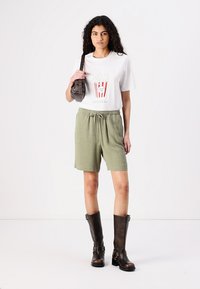Witte t-shirt met popcornafbeelding, olijfgroene shorts met elastische tailleband, gecombineerd met kniehoge bruine leren laarzen en een kleine tas.