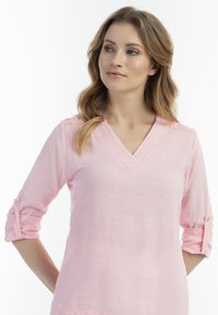 Lichtroze linnen blouse met een V-hals, driekwartmouwen en decoratieve kantaccenten op de schouders. Zachte textuur.
