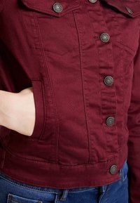 Main dans la poche d'une veste en denim bordeaux ajustée avec des boutons en métal, portée sur un jean bleu, montrant le devant de la veste et les détails de couture.