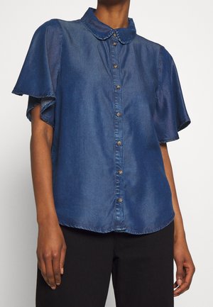 Person iført en blå denim kortærmet bluse med flagrende ærmer, knaplukning foran og afrundet krave, kombineret med sorte bukser.