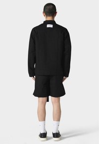 Black jacket and shorts in a sleek, minimal design. Made of smooth fabric with a straight cut. Worn with white socks and black shoes.