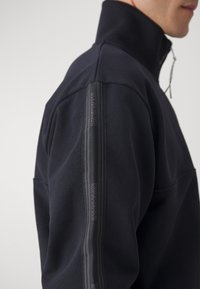 Homme portant une veste noire zippée à col montant avec une bande verticale marquée sur la manche, sur un fond clair uni.