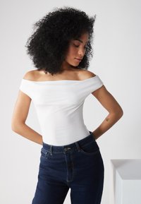 Witte off-the-shoulder top van gladde stof, gecombineerd met donkerblauwe high-waisted jeans met subtiele stiksels en klassieke hardware.
