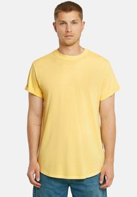 T-shirt giallo a maniche corte in tessuto leggero con scollatura rotonda e maniche arrotolate. Presenta un logo discreto sul davanti.